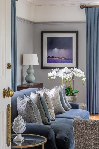 Grey velvet sofa with bespoke cushions, blue wool curtains and potted iris in the background.