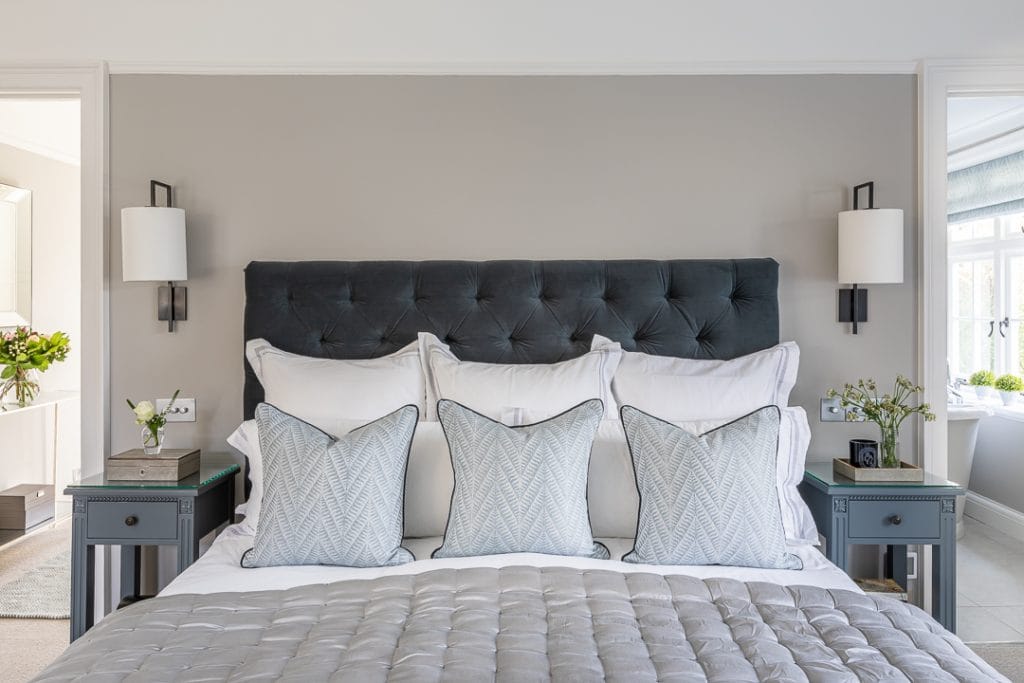 Upholstered dark grey button back headboard with bronze wall lights and cream silk shades, grey silk quilted bedspread and blue patterned cushions.