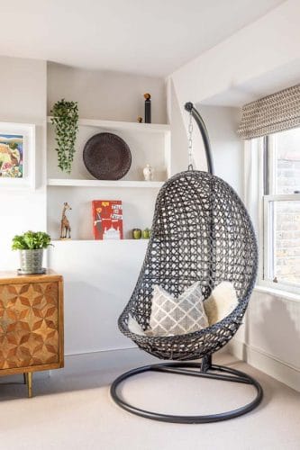 Black wicker egg shaped hanging chair in playroom with marquetry side board and shelves displaying books and toys.