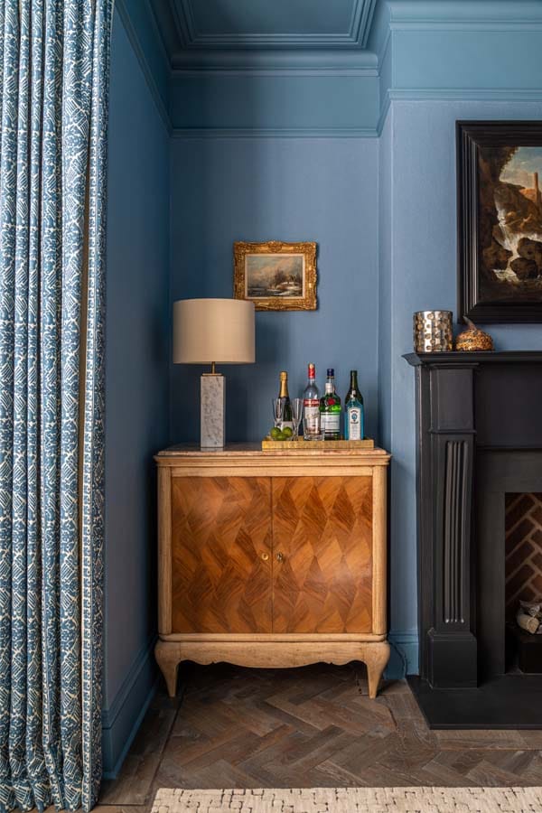 Antique inlaid drinks cabinet with marble lamp and drinks tray. Blue linen wallpaper and hand printed curtains.